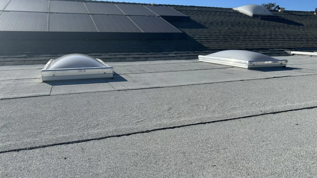 A flat roof with two skylights in the foreground and solar panels on a sloped roof behind them.
